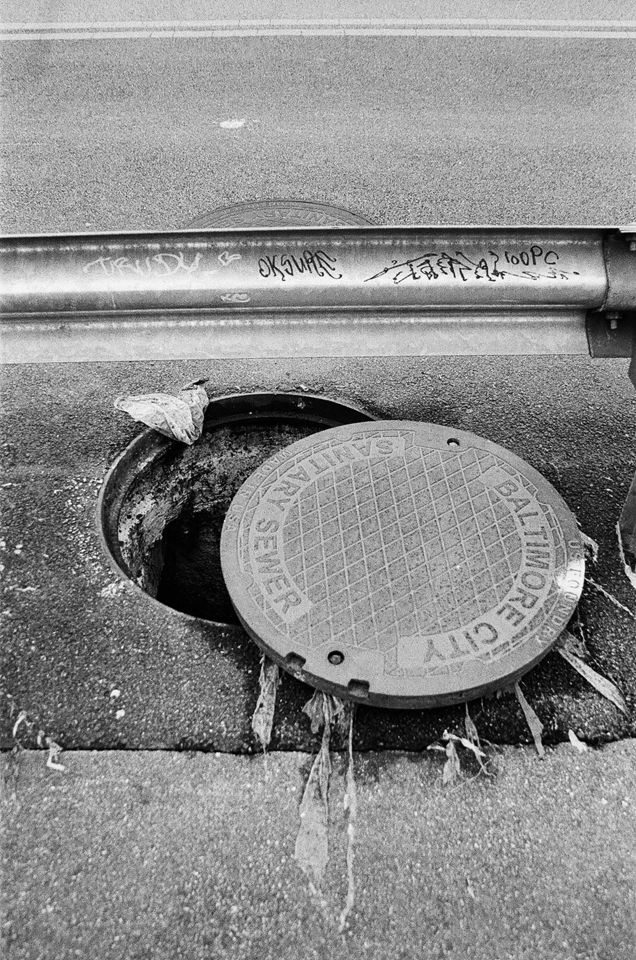 A city sewer cover is shown popped out of the ground next to a street and guard rail.