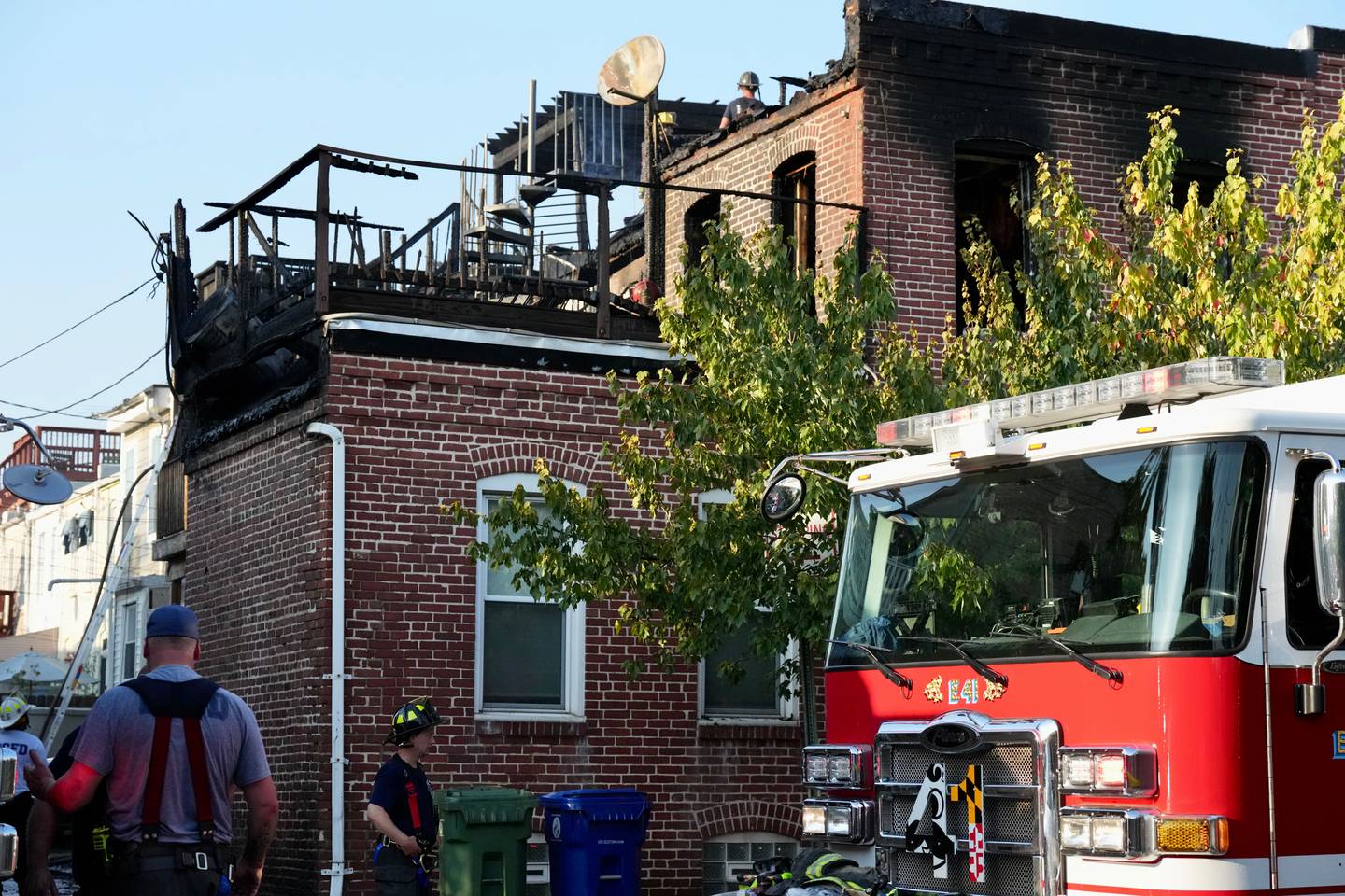 A firefighter stands on the roof of a burned rowhome. A burned deck and staircase are visible. In the foreground is a Baltimore Fire Department truck.