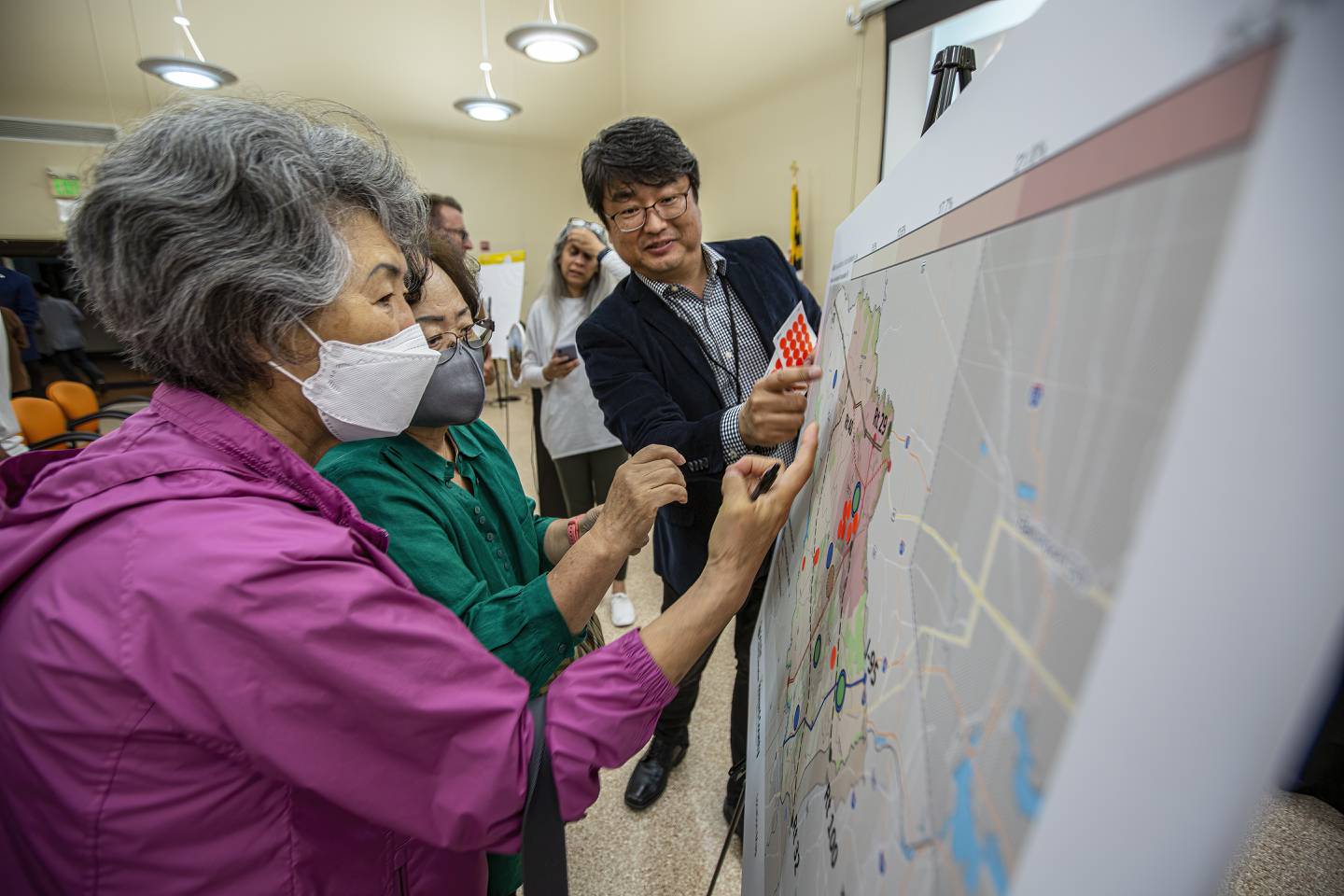 In the foreground, an Asian woman with grey hair looks at a map on a display stand.