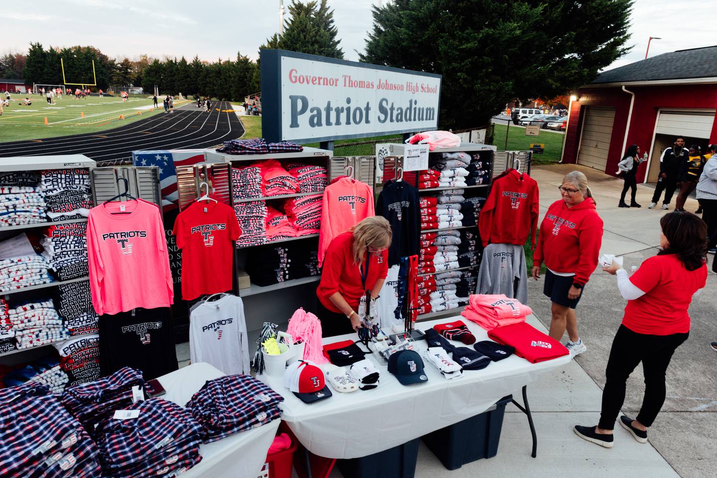 Thomas Johnson Patriots merchandise table attendants, Sherry Apperson, left, and Dawn Buckley, center-right, sell TJ apparel following the final regular season game of the first varsity flag football season at Frederick County Public Schools, on Thursday, Oct. 26, 2023 in Frederick, MD. The Baltimore Ravens helped fund the FCPS girls flag football pilot program this year. (Wesley Lapointe / for the Baltimore Banner)