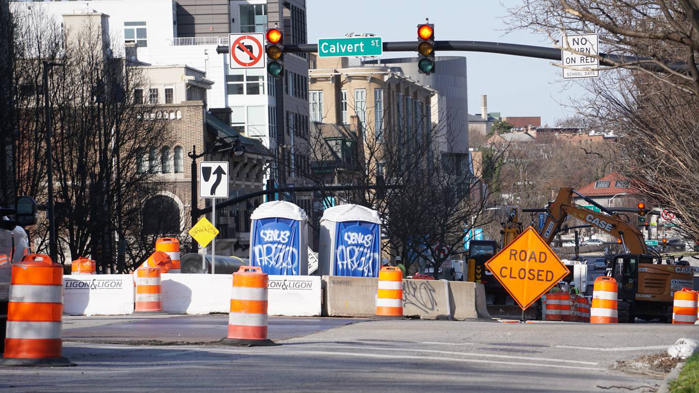 An orange roadwork sign that reads "road closed" blocks off a section of an intersection. A green road side that says "Calvert St" hangs next to a red stop light. There are some orange roadwork barrels and white traffic barriers.