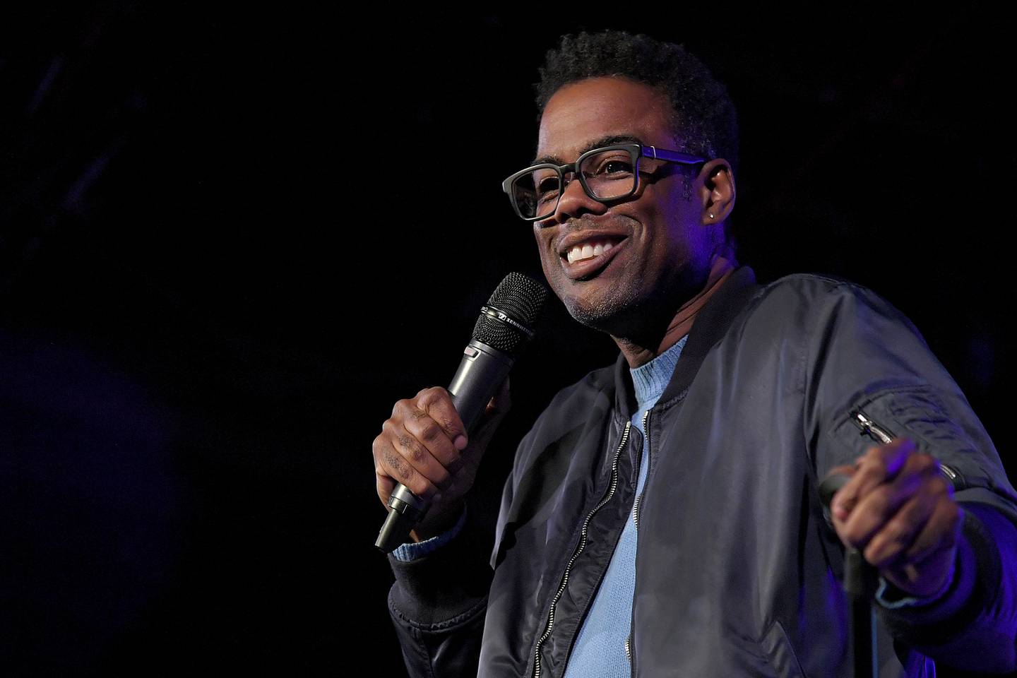 Comedian Chris Rock holds a microphone while performing. He's wearing glasses, a dark jacket and blue shirt.