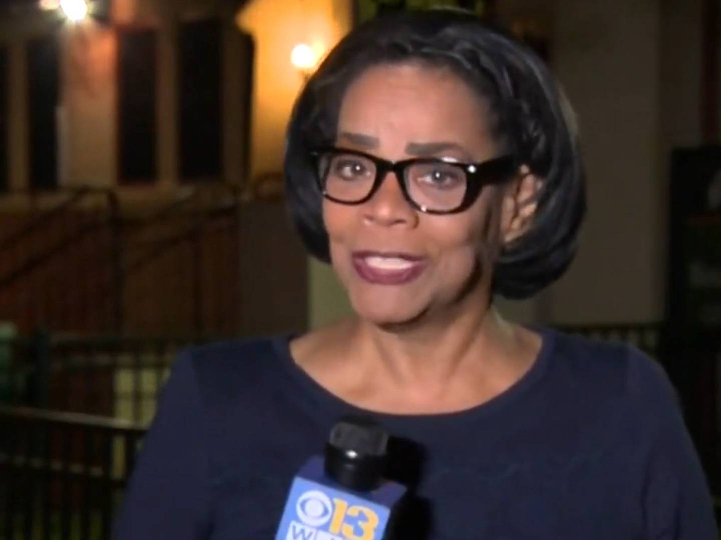 A woman wearing glasses holds a microphone and speaks to the camera. A screen grab of a news broadcast.