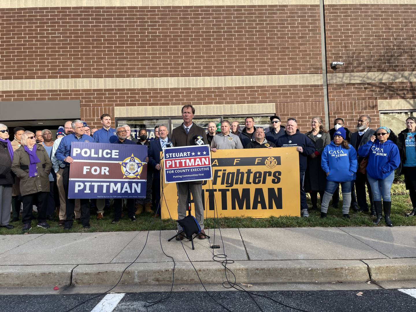 Anne Arundel County Executive Steuart Pittman stands at a lectern speaking. Behind him are supporters from police and fire fighter unions.