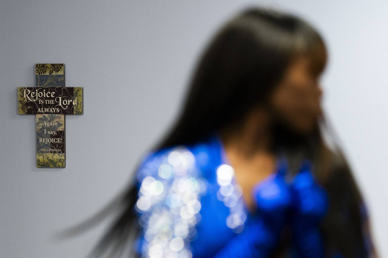 A model walks in a fashion show for Donna Bruce. Behind her, a cross hangs on the wall that says "Rejoice in the Lord always!". Bruce is very invested in her faith.