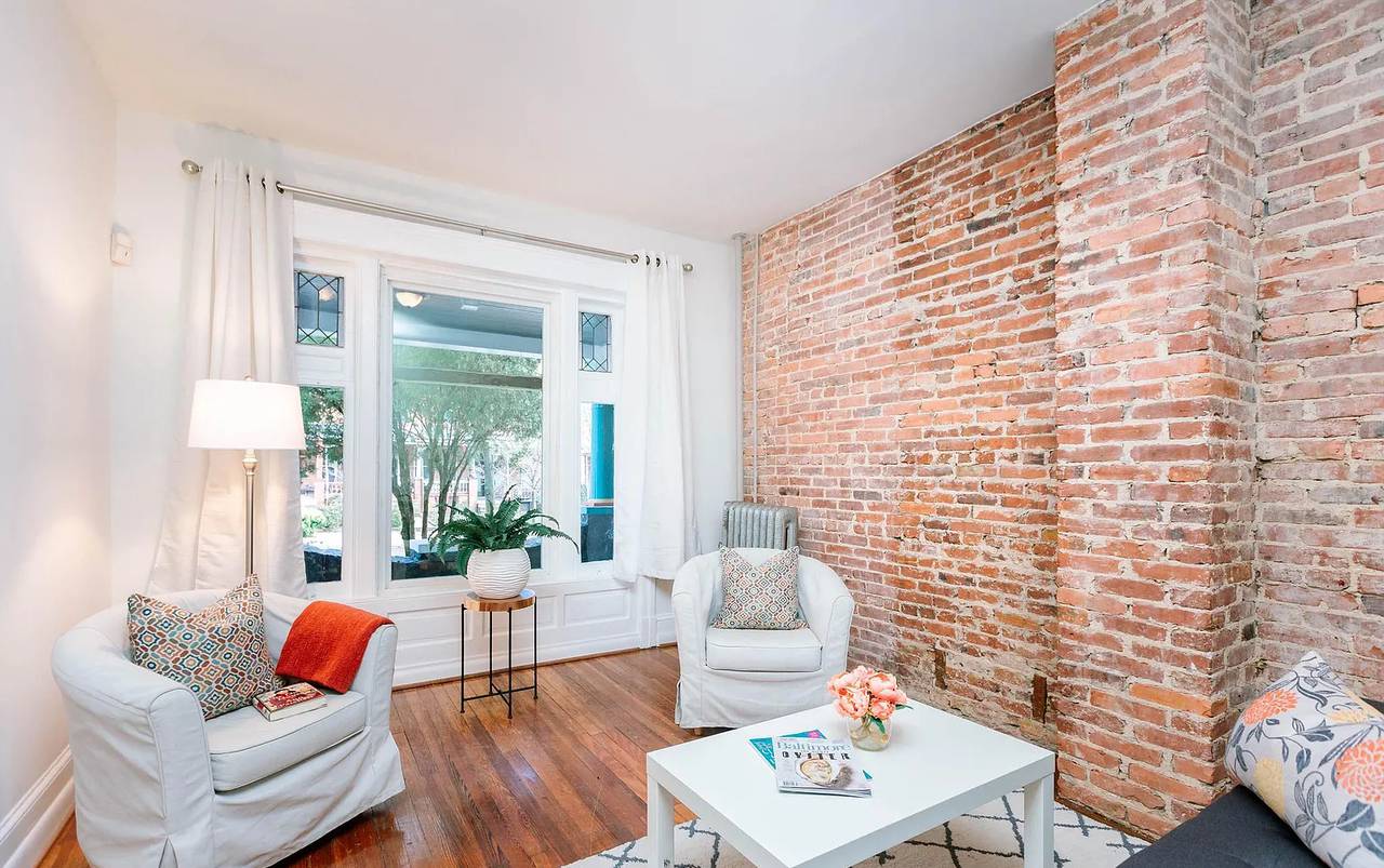 A living room with an exposed brick wall, wood floors, white furniture and a large window.