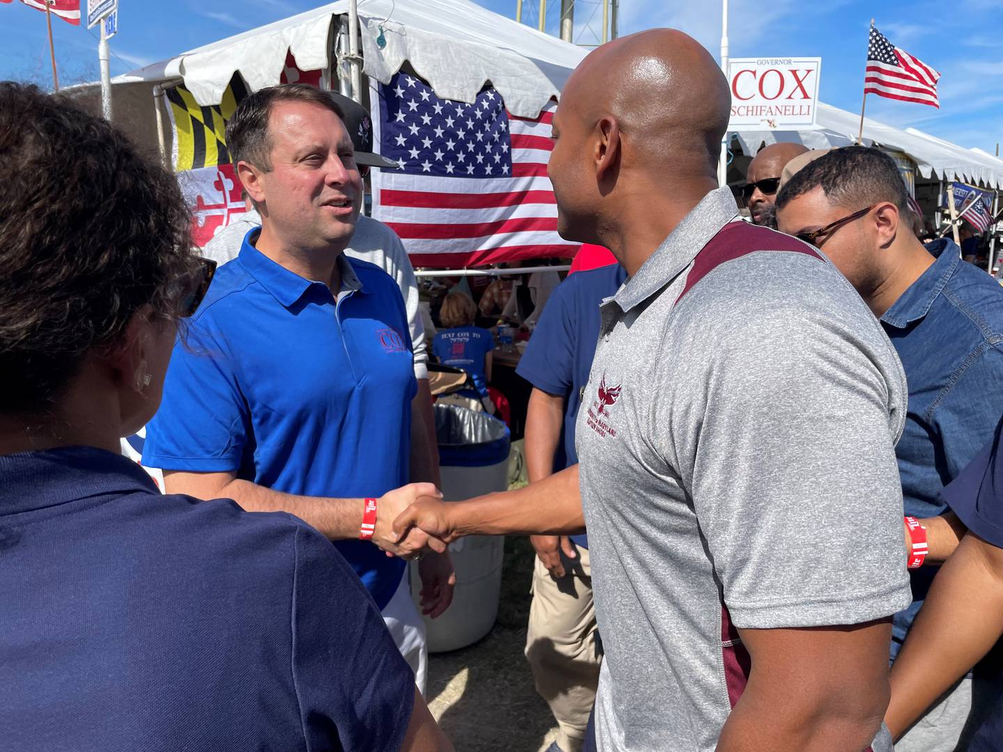 Dan Cox, in a blue shirt, shakes hands with Wes Moore, in a gray shirt, in front of a tent with Maryland and United States flags.