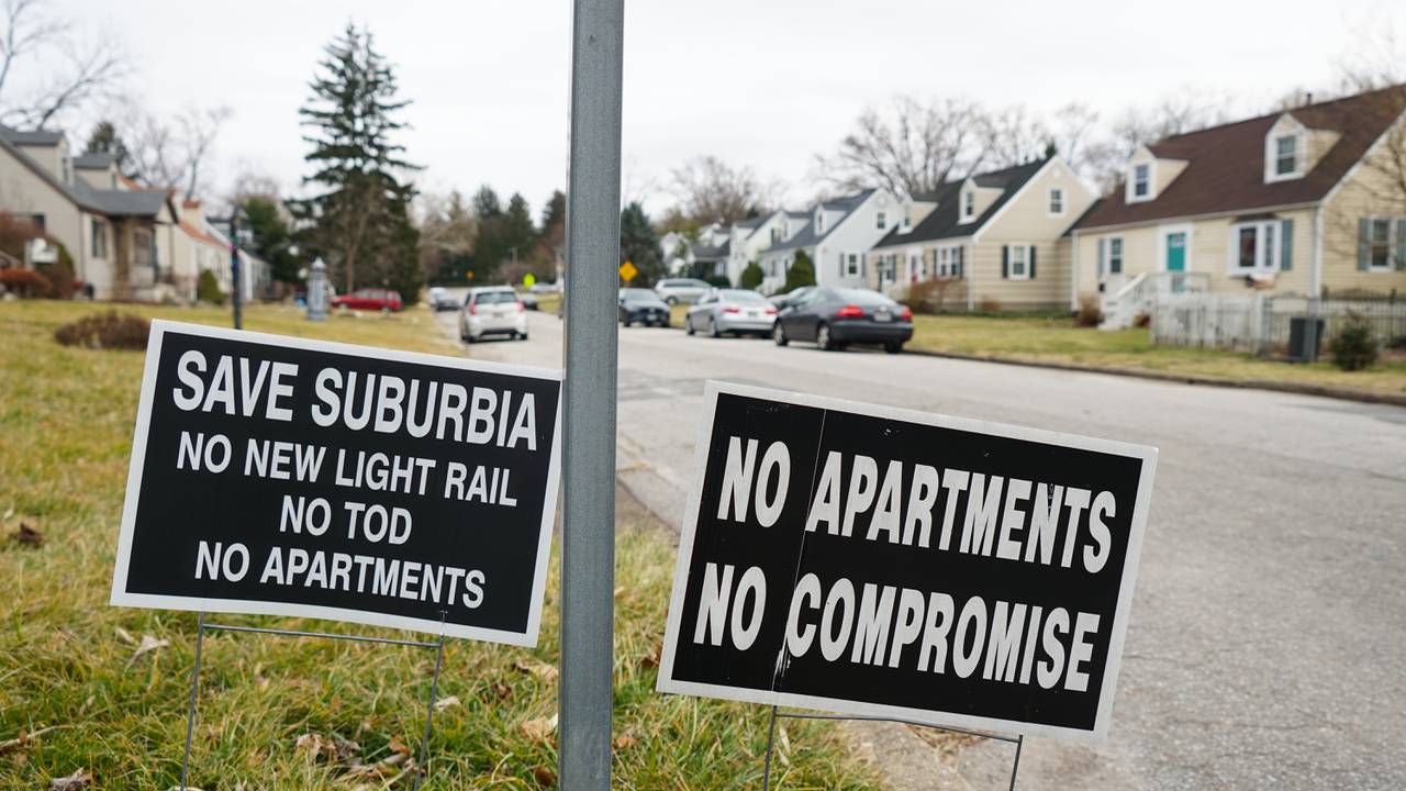 Two black and white yard signs, one that reads "save suburbia, no new light rail, no TOD, no apartments" and the other "no apartments, no compromise" are staked into the grass in front of a suburban street with cars and single family homes in the background.