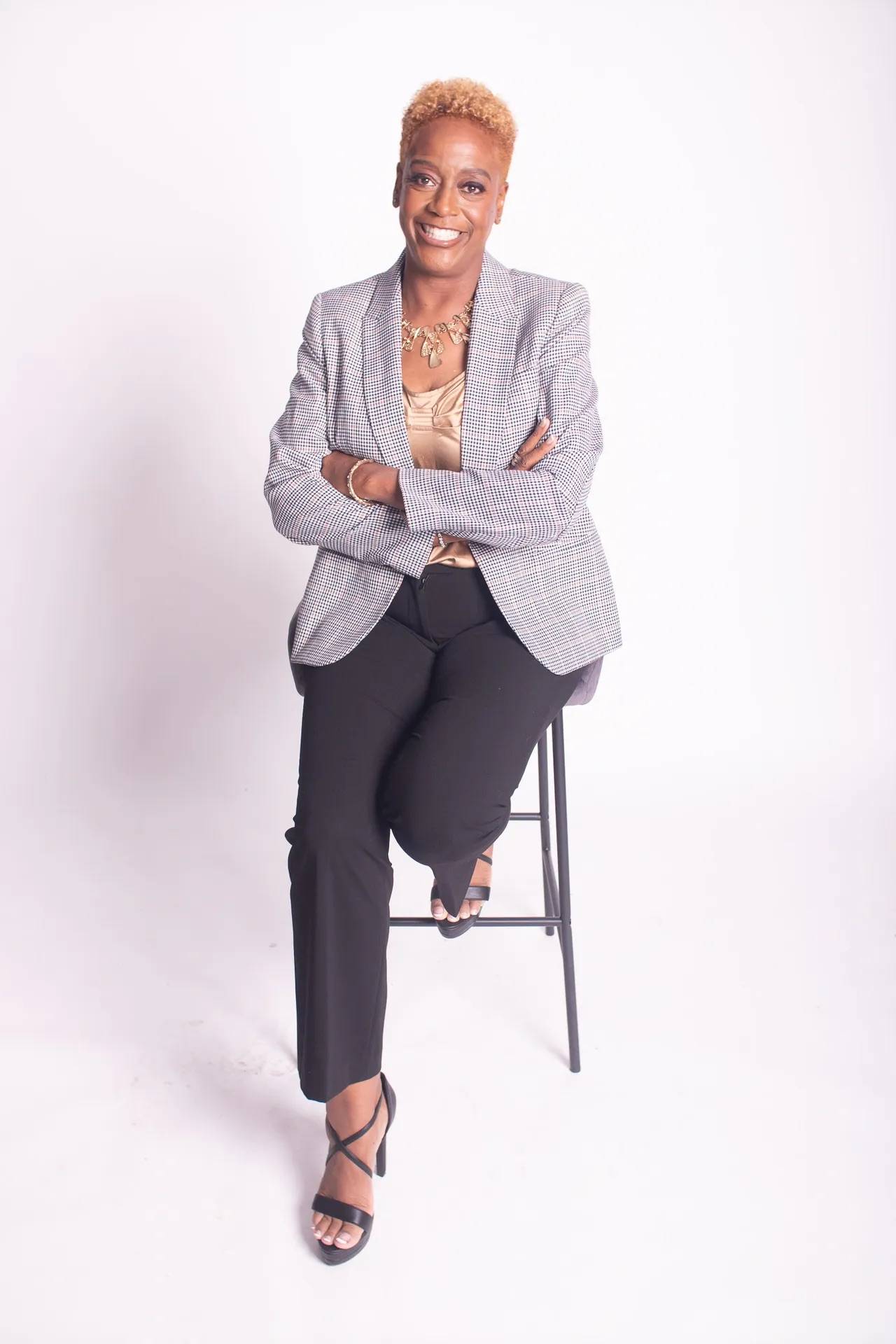 A photo of Robyn A. Christian wearing a great suit jacket and black pants, sitting on a stool in a studio with a white background.