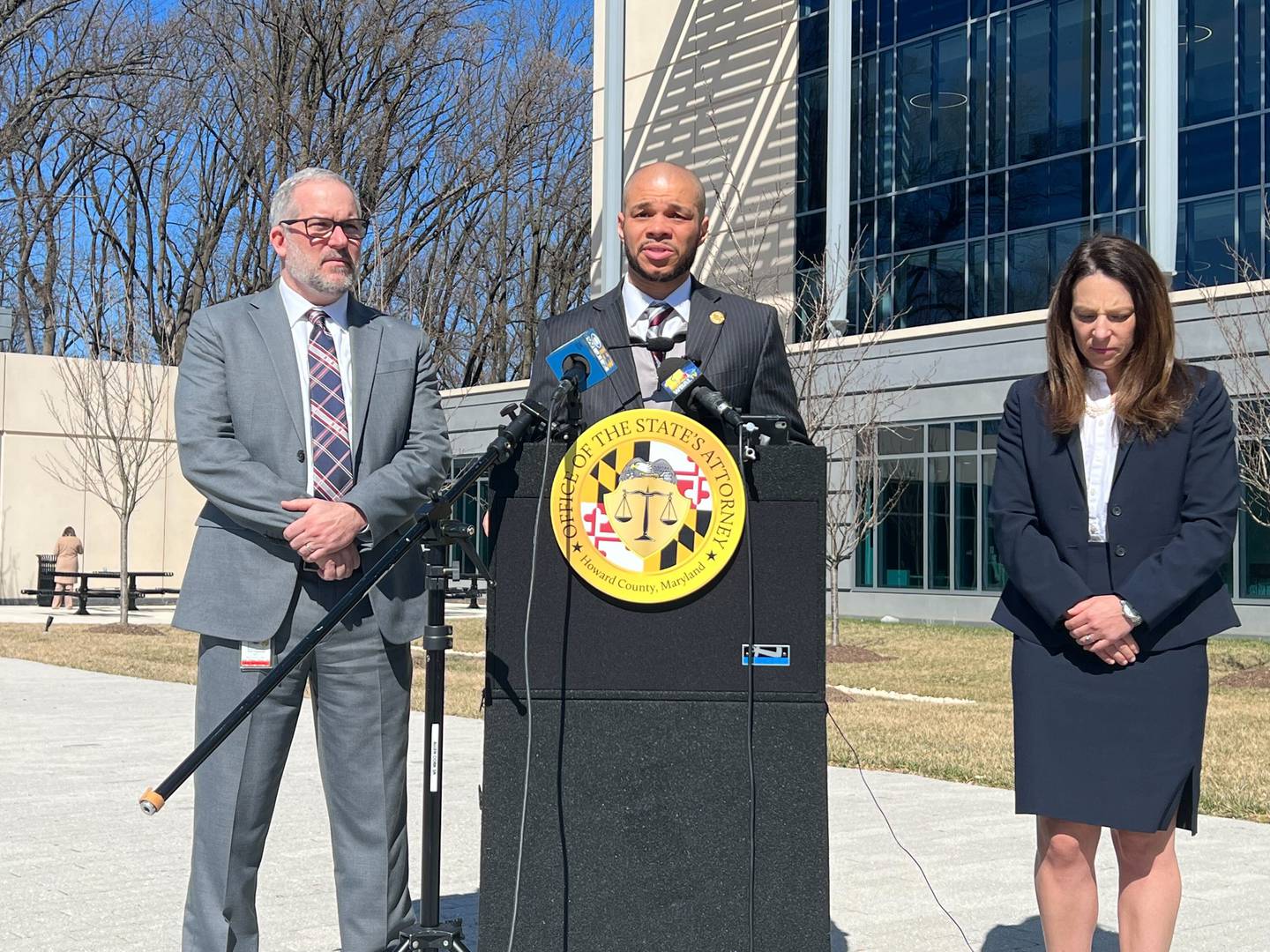 A man in a dark suit stand at a lectern in front of the Howard County courthouse. On his right is a man in a gray suit and on his left is a woman in navy suit.
