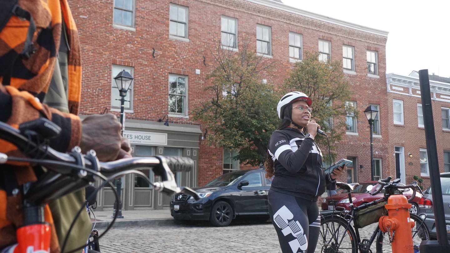 A woman wearing a bike helmet speaks into a microphone on a city corner.