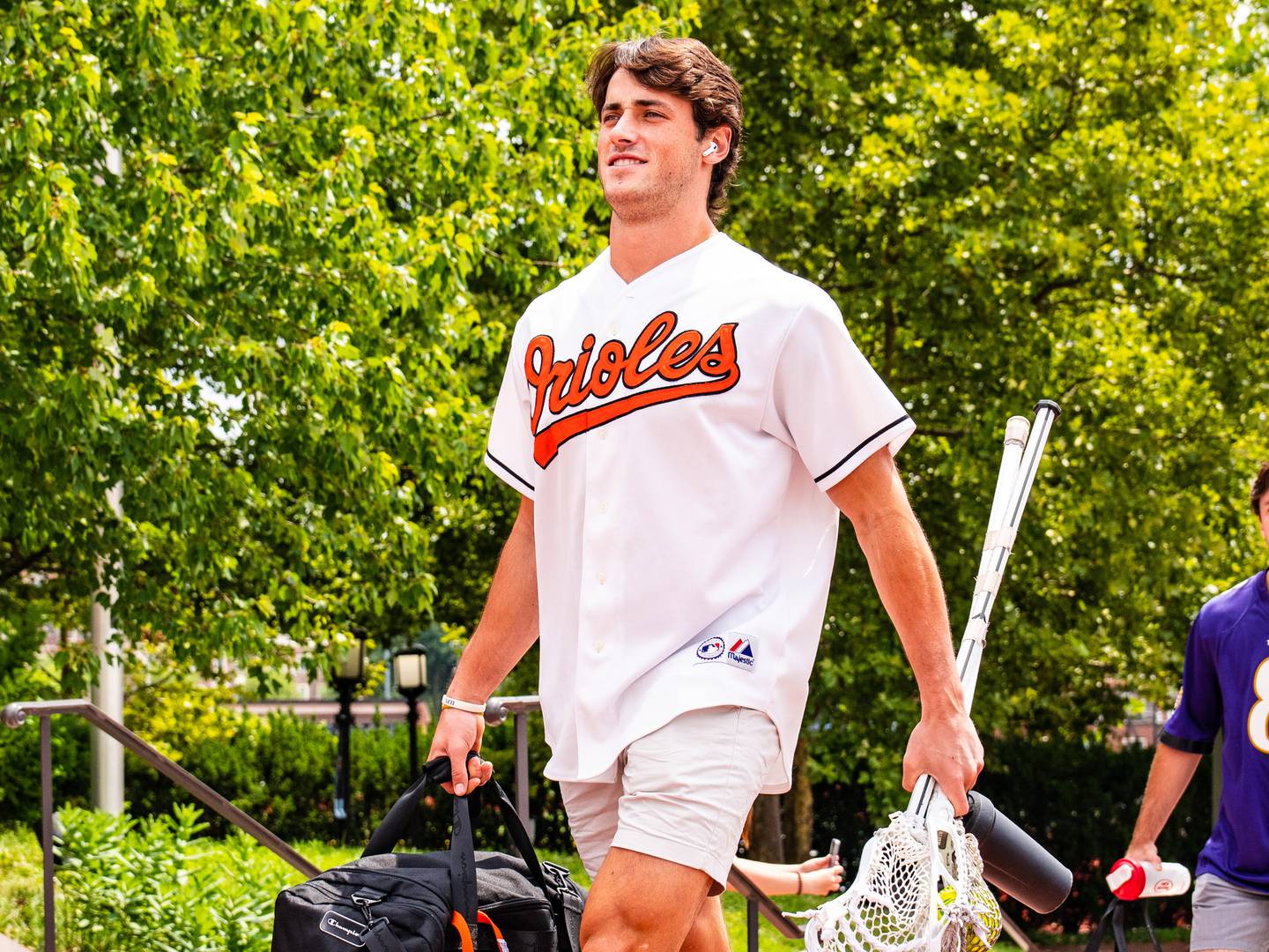 This photo shoes a lacrosse player holding equipment and wearing and Orioles jersey walking along a brick-lined path.