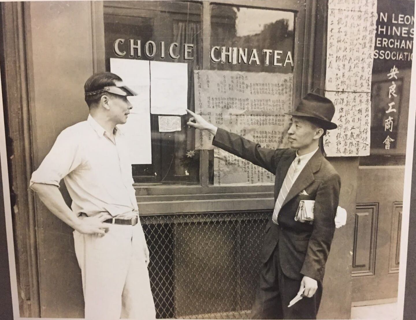 In this sepia-toned photo, two men stand outside a storefront that reads "Choice China Tea." One man points at papers posted in the window. Chinese calligraphy hangs in strips near