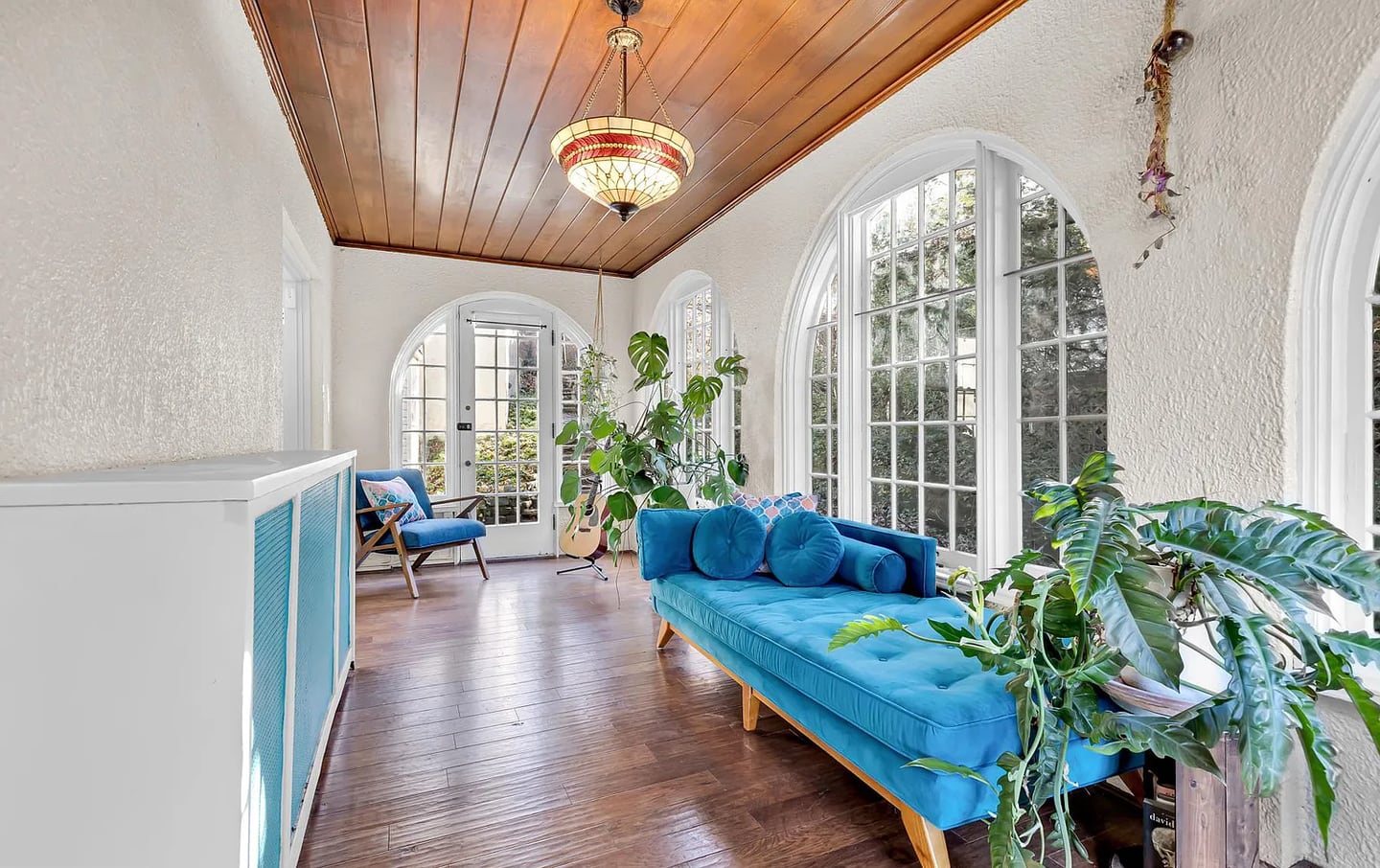 A bright sunroom with arched windows, dark wooden floors, a wood-stained ceiling and bright blue furniture.