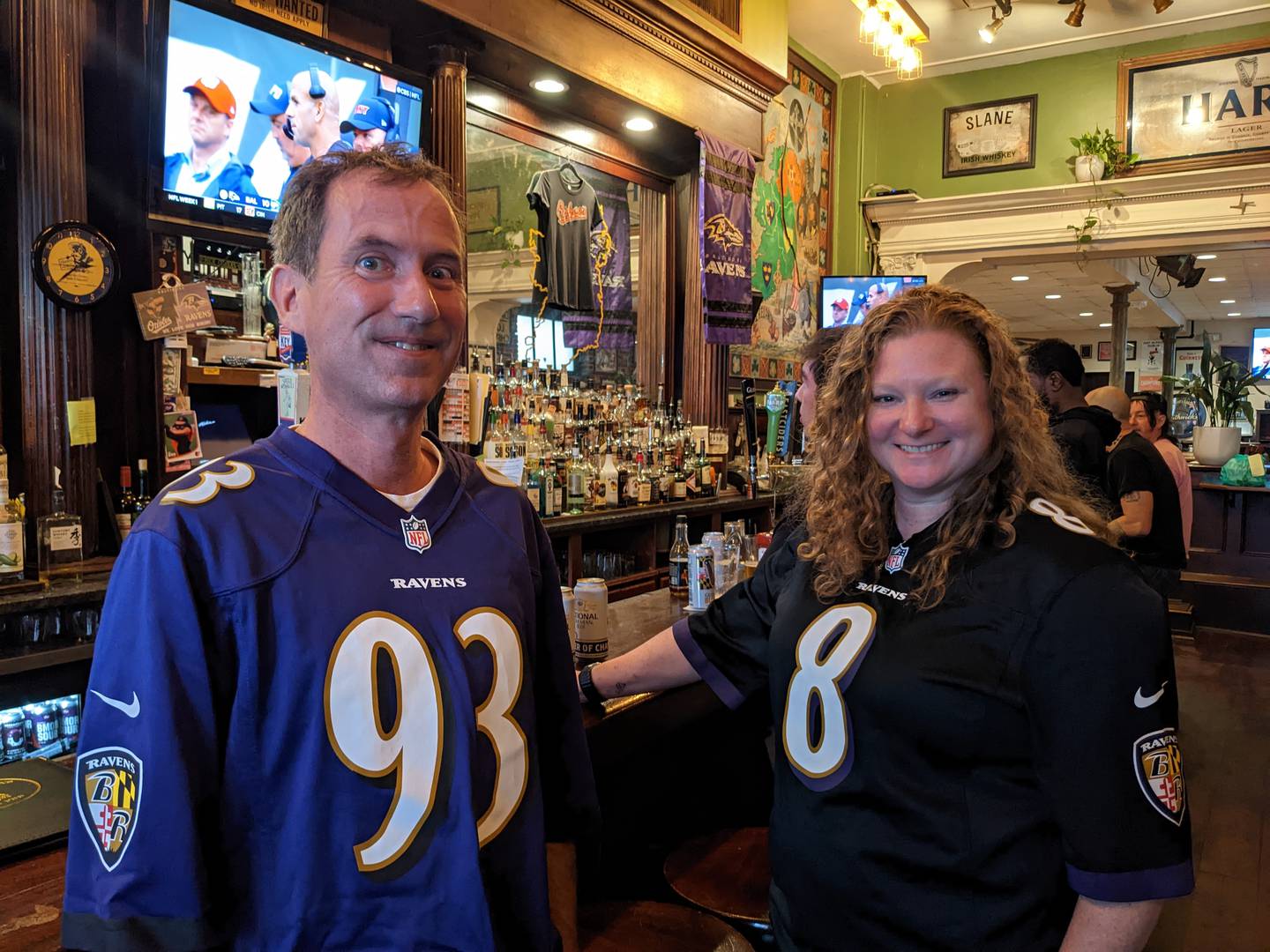A man and a woman, both wearing purple Ravens jerseys, stand in front of a bar playing football on the television.