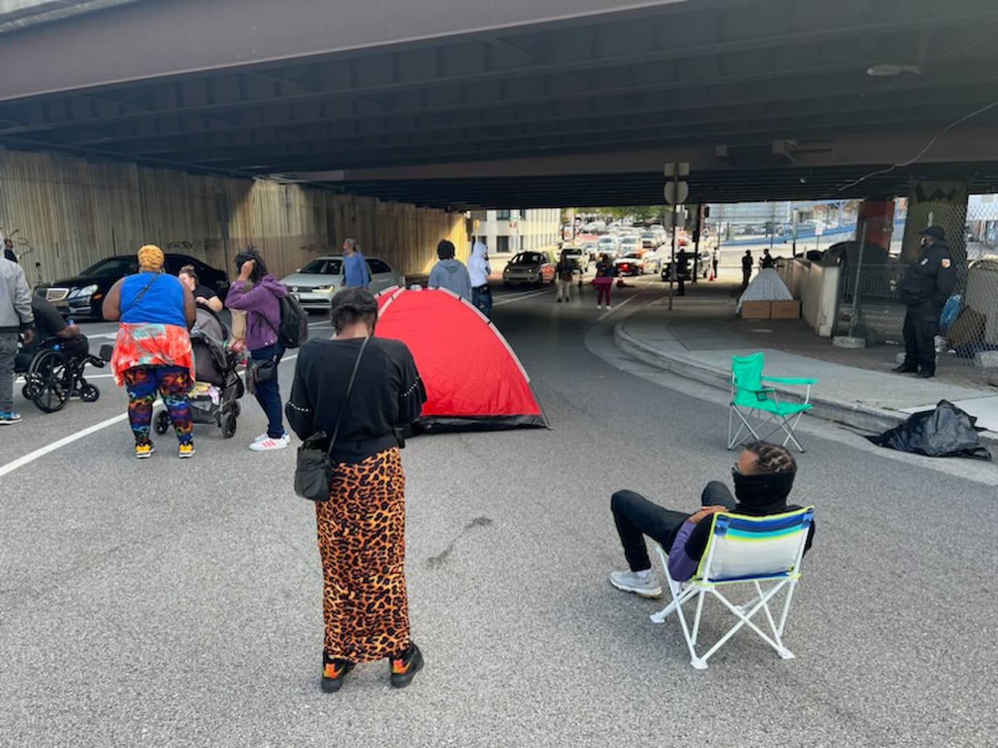 People standing in the road blocking traffic. One person is sitting in a chair. Tents are visible in the background.