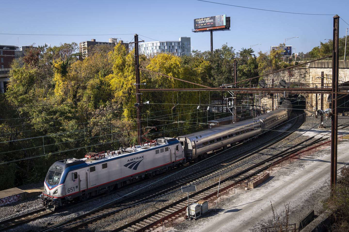 A silver and blue Amtrak train emerges from a tunnel. In the background trees and a small piece of the Baltimore skyline are visible.