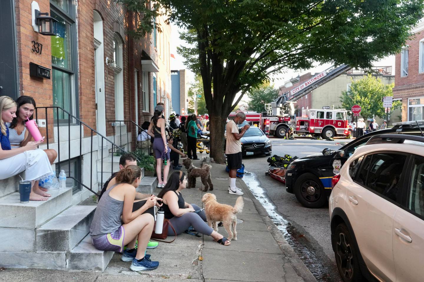 A group of people sit on stoops and the sidewalk as fire truck are seen in the background.