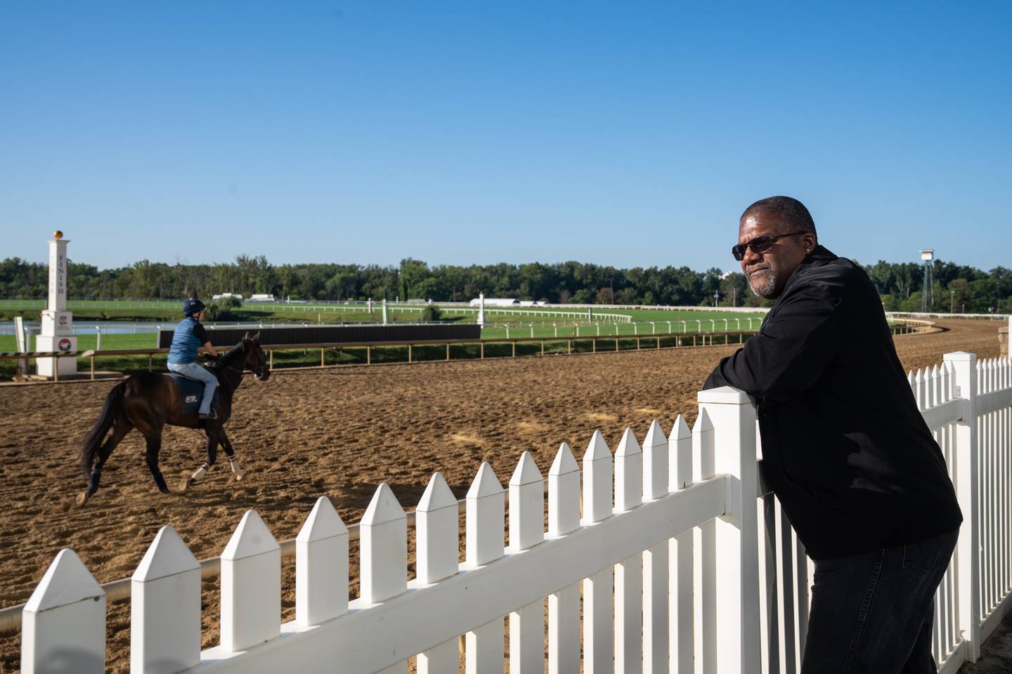 Jerome Aiken leans on a white fence as he watches jockeys and horses go by on the racetrack.