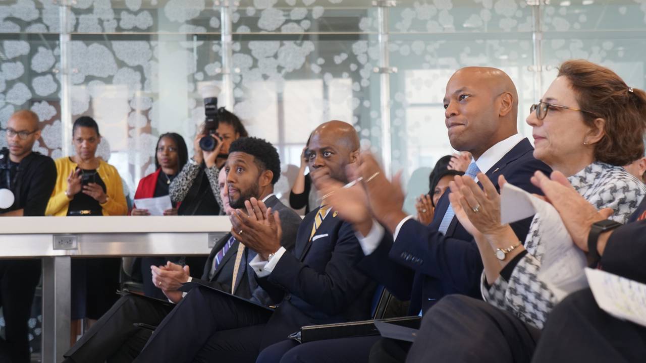 Baltimore Mayor Brandon Scott, Coppin State University President Dr. Anthony Jenkins, Gov. Wes Moore and U.S. DOT Deputy Director Polly Trottenberg sit in a row chairs and clap.