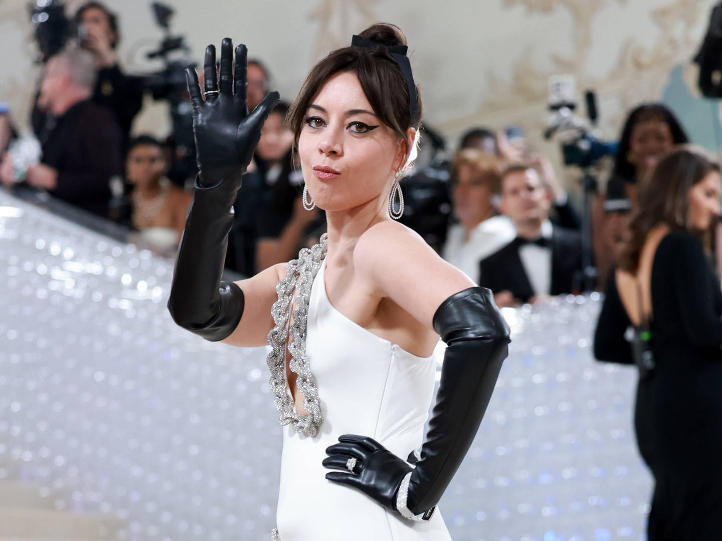 Actress Aubrey Plaza waves at the crowd at the entrance to the 2023 Met Gala  while wearing black gloves and a white gown.