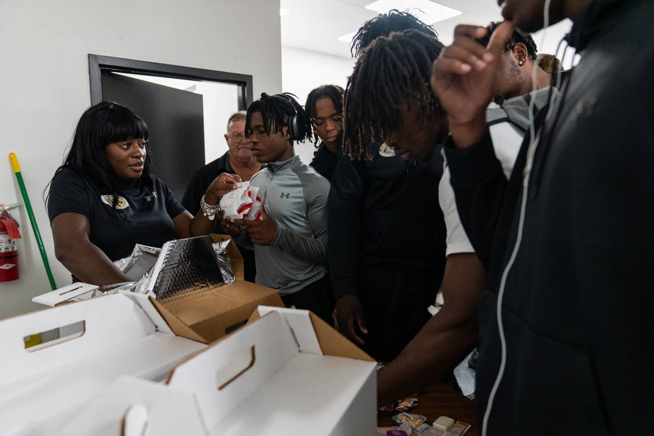 Travel Coordinator Ericka Allen, left, hands out packaged Chick-fil-A sandwiches to a line of players.