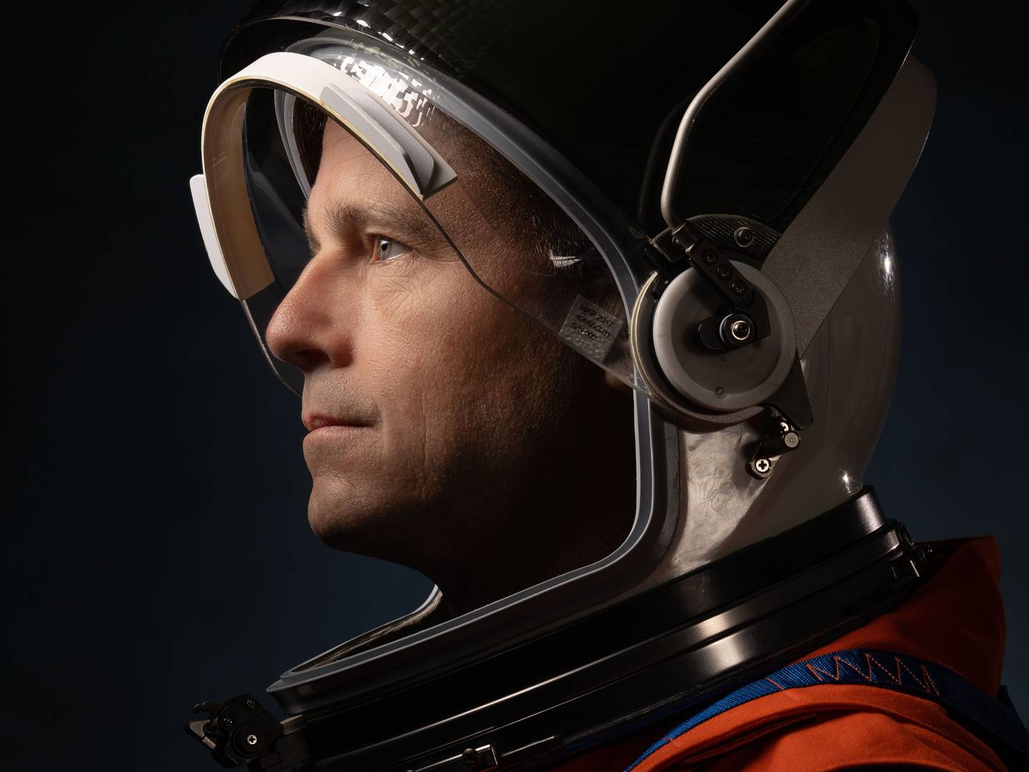A middle-aged caucasian man wears an astronaut helmet with a visor lifted up and looks off to the left in a portrait against a black background.