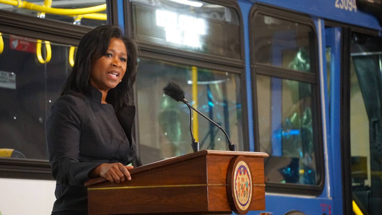 A woman stands in front of a brown podium and speaks into a microphone. There is a blue and white bus in the background with a bright LED sign that reads, "let's clean the air."