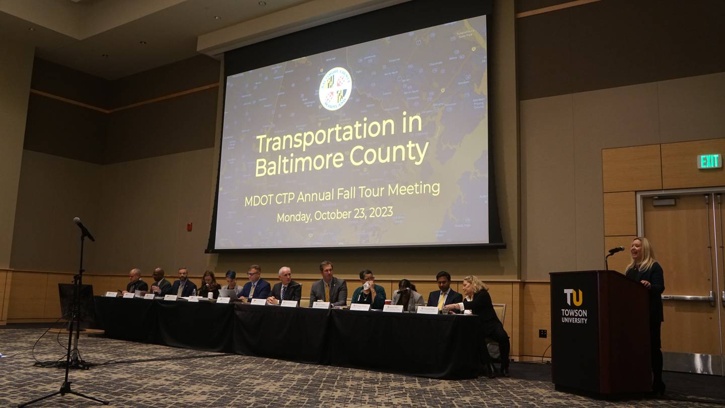 Men and women sit at a long panel-style table in a large room underneath a projector screen holding a presentation called "Transportation in Baltimore County."