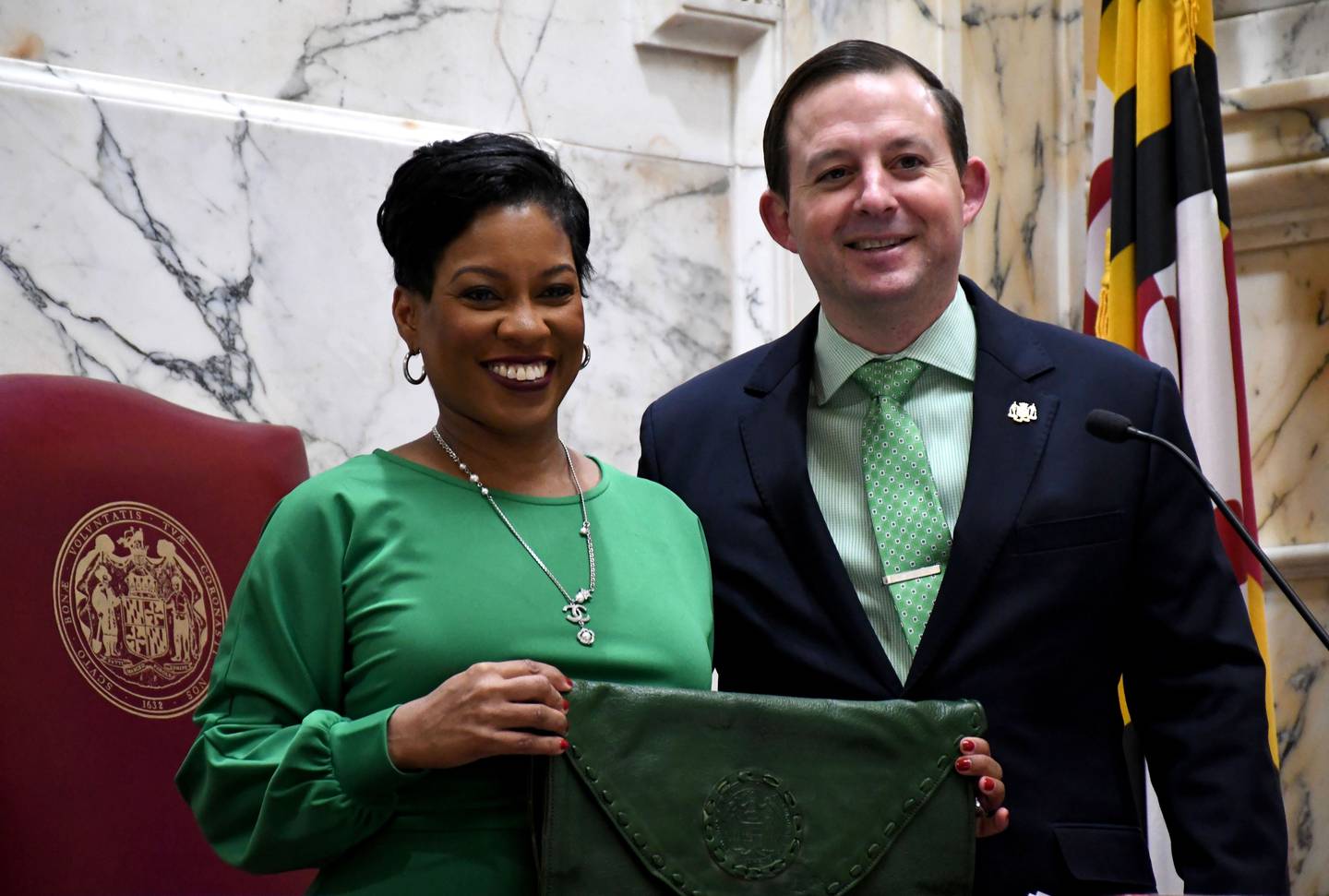 A woman wearing a green dress smiles and holds a green leather bag. Next to her is a man in a dark suit with a green shirt and tie. A Maryland flag and marbled wall is behind them.