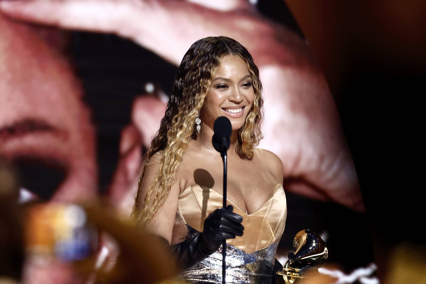 Beyoncé smiles as she accepts a Grammy Award wearing a gown and standing in front of a microphone.