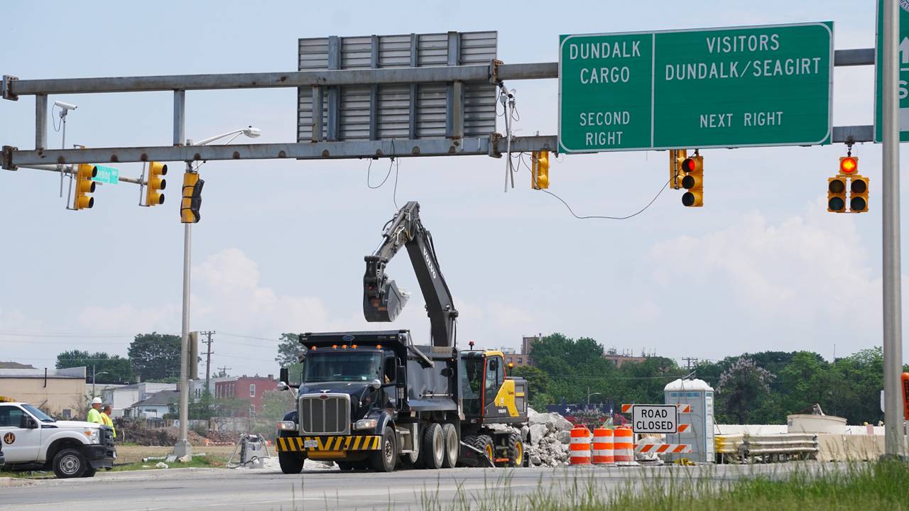 A crane is lifting debris into a dump truck underneath a road sign that indicates directions toward the Port of Baltimore.