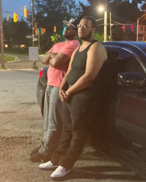Photograph of two African American men leaning against a car at dusk.