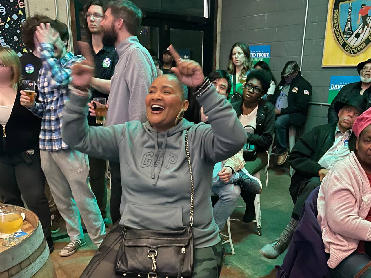 A woman wearing a grey sweatshirt and sitting on a stool in a brewery smiles and raises her arms above her head while watching a debate.