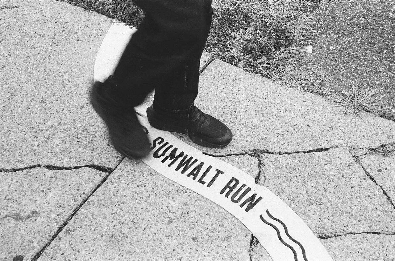 The feet of a man are shown walking on a cracked city sidewalk over a marking that reads "Sumwalt Run".
