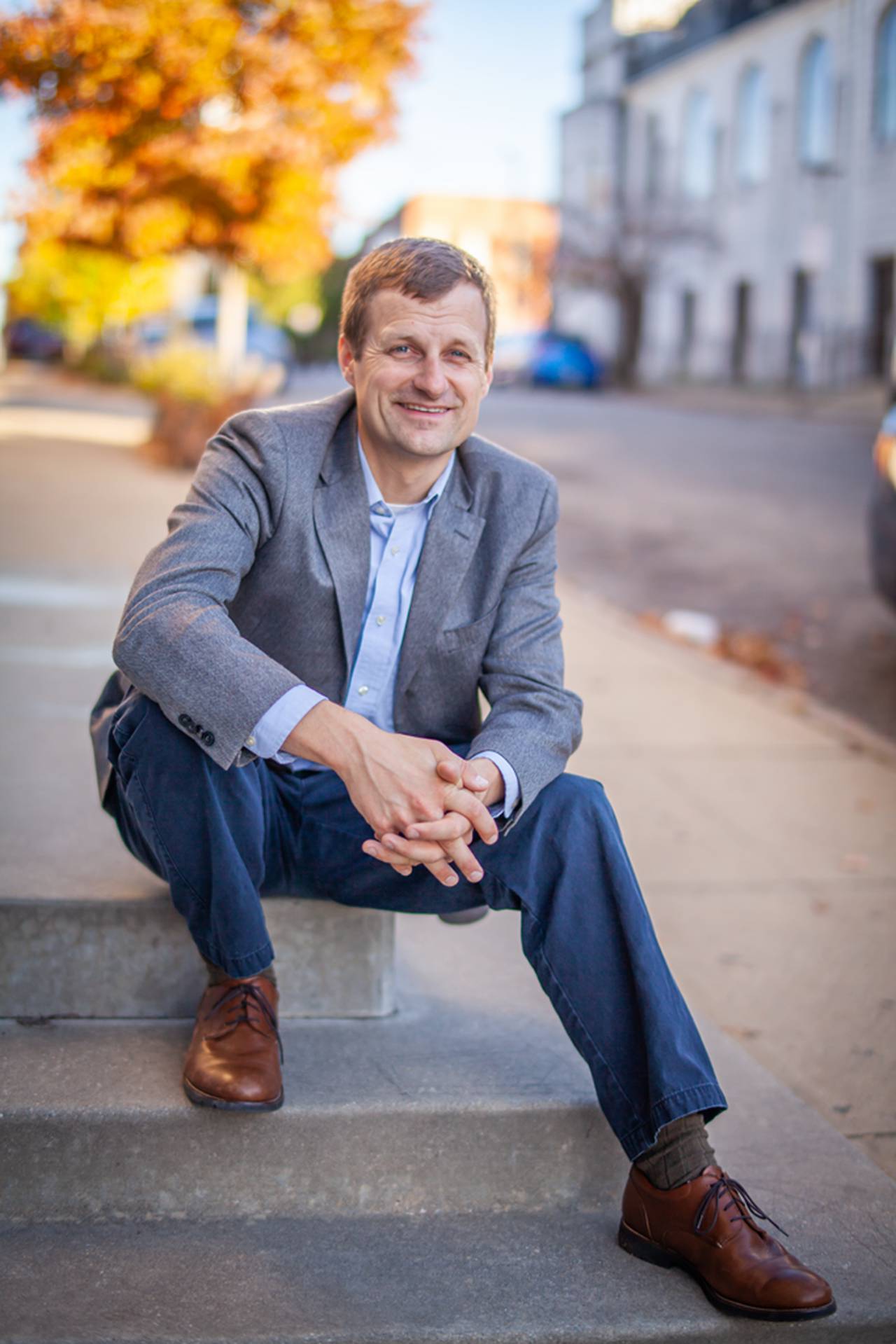 A photo of Mark Parker sitting on steps wearing a gray suit jacket, a light blue shirt, dark blue slacks, and brown shoes.