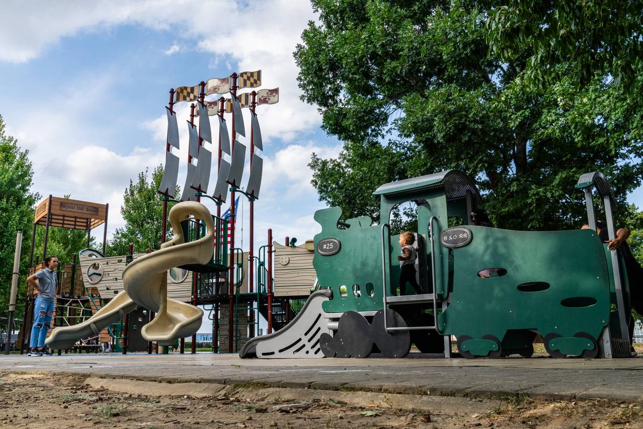 A wide shot of Federal Hill Park Playground's three history-themed structures: (from left) Signal Hill Tower, The Federalist ship and the B&O Railroad. A running child is framed through the railroad car opening.