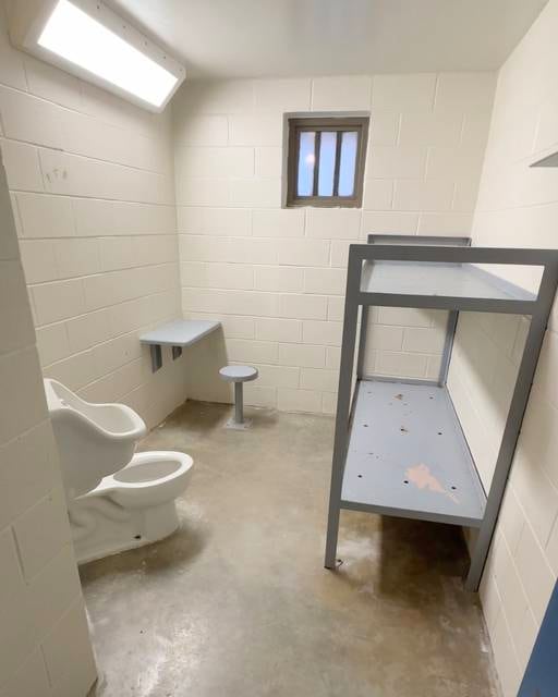 Photograph of the empty interior of a prison cell. On the left are a urinal, toilet and small desk with stool. On the right is a double-decker meta bunk bed.