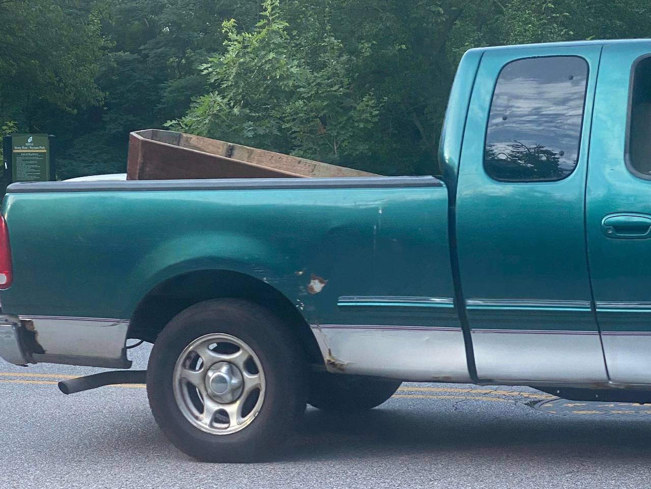 A teal pickup truck carrying an empty coffin in the bed of the truck drives away from a park.