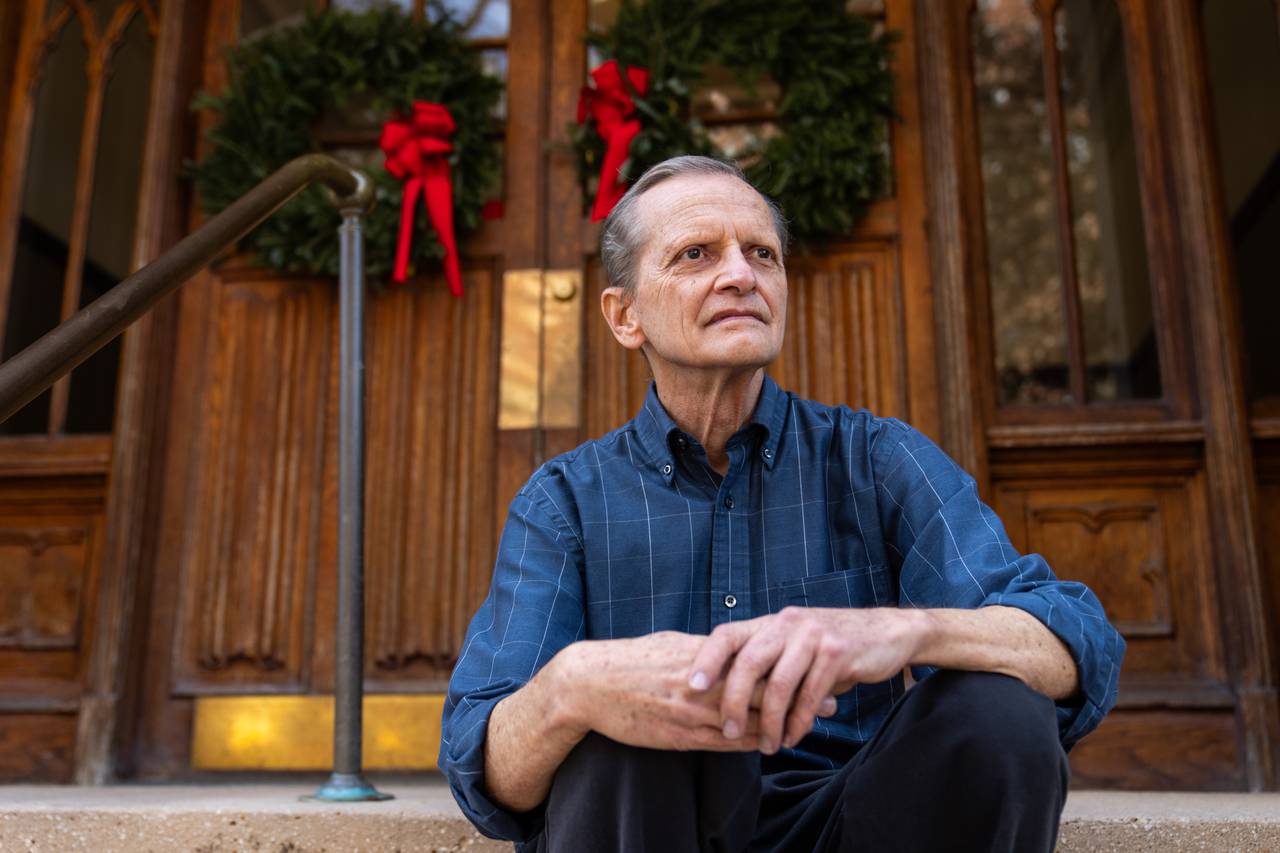 Richard Oloizia poses for a portrait, looking away from the camera and sitting with his hands clasped in front of Tudor Arms Apartments' wooden doors, which are decorated with wreaths.