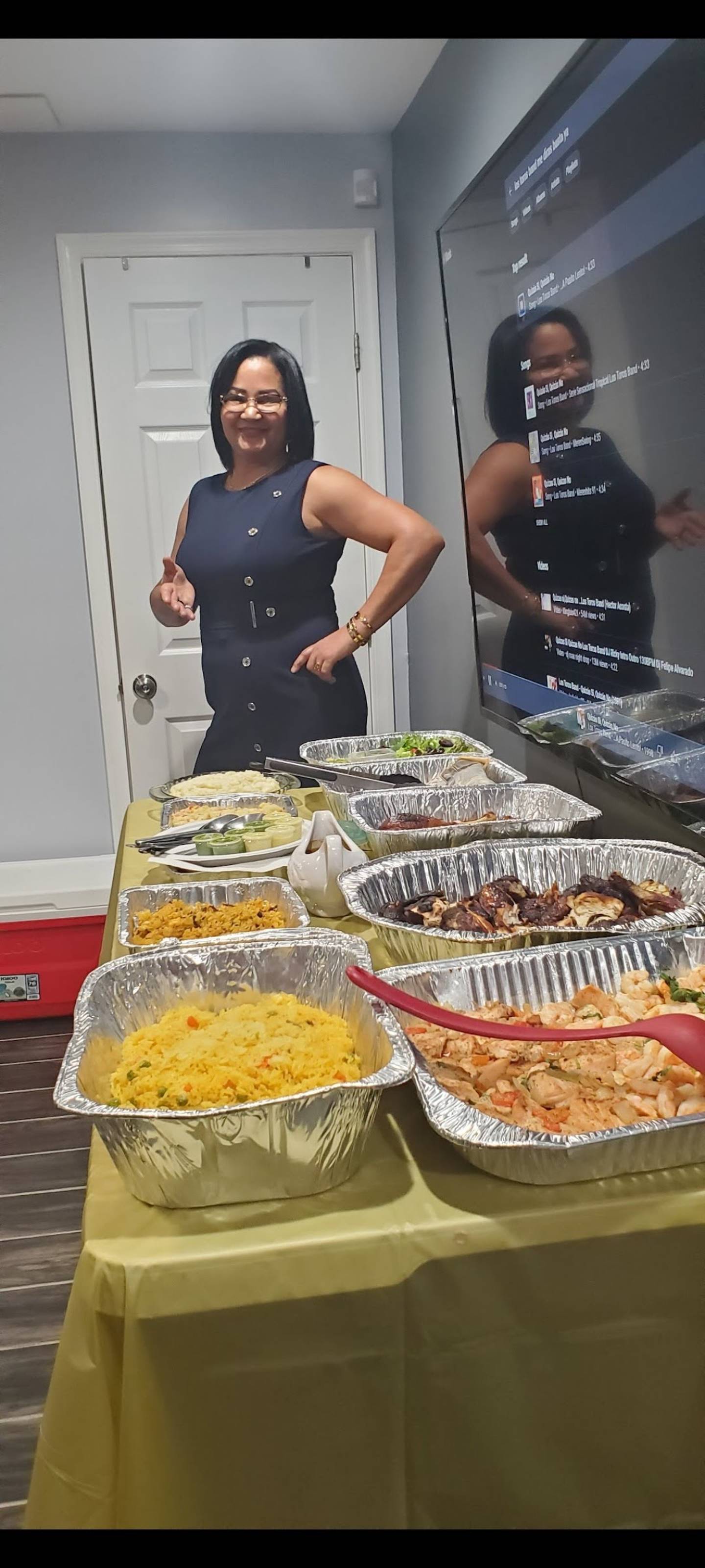A woman with glasses and a dark dress gives a thumbs up. On a table in front of her is a variety of food, served in large aluminum pans.