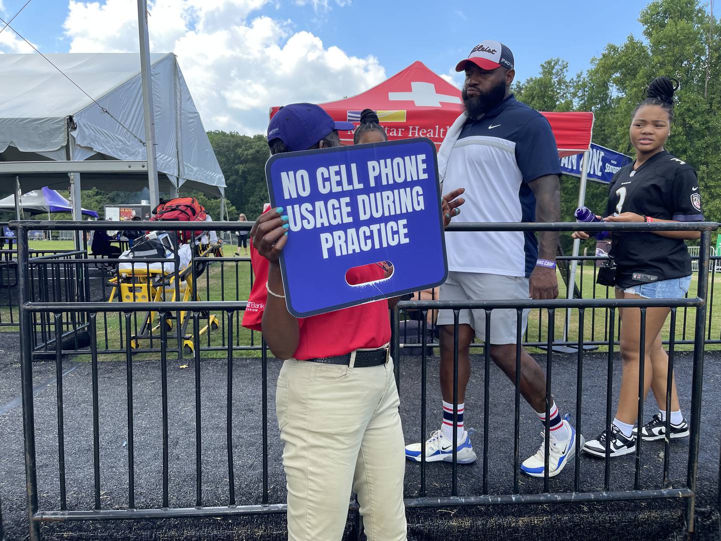 An employee from S.A.F.E., the security company, with a sign that says "NoCell Phone Usage During Practice."